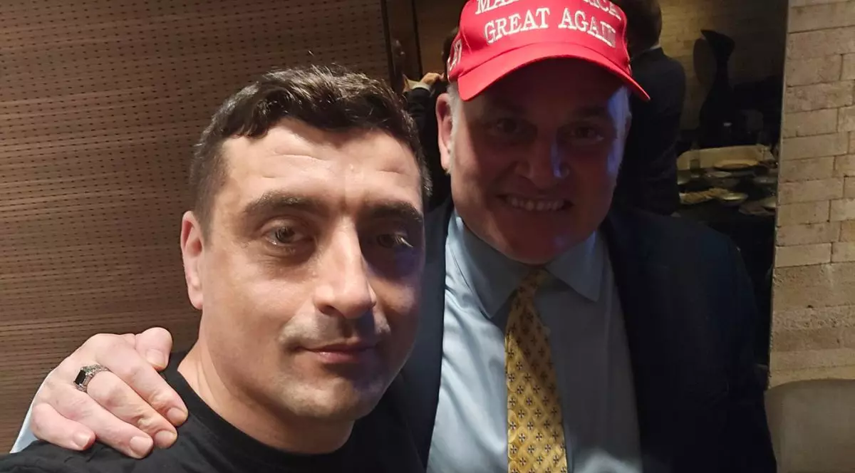 George Simion, într-un tricou negru, ținut de după gât de Brian Brown, susținător MAGA, îmbrăcat într-un costum bleumarin cu o cămașă bleu și o cravată aurie, cu o șapcă roșie cu textul „MAGA”