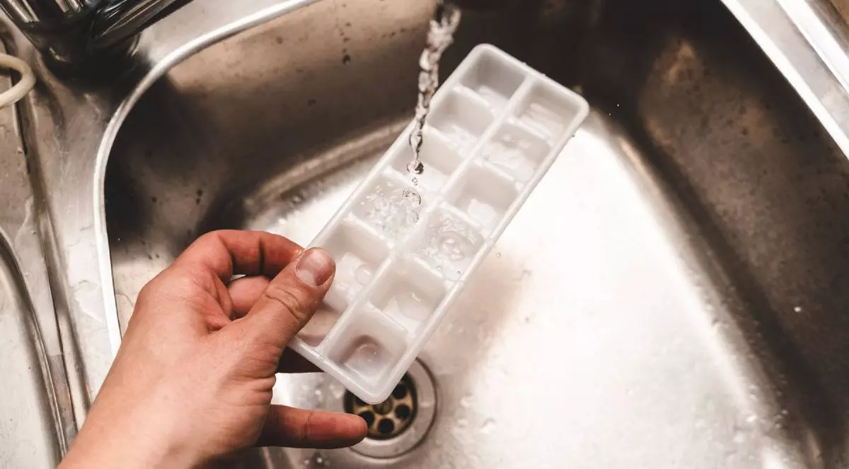 Mâna unei femei, care ține o formă de cuburi de gheață, în timp ce apa umple formele într-o chiuvetă