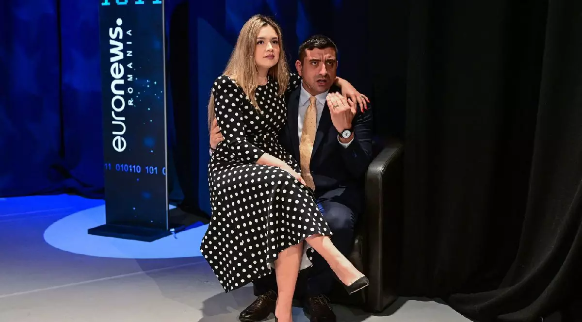George Simion și soția sa Ilinca în timpul unei dezbateri împotriva adversaruluiNicușor Dan (nu apare în fotografie) organizată de Euronews România la București, 8 mai 2025. Ilinca, îmbrăcată într-o rochie neagră cu buline albe, stă ăn brațele lui George Simion, care este îmbrăcat într-un costum închis la culoare, cămașă albă și cravată aurie, și gesticulează.