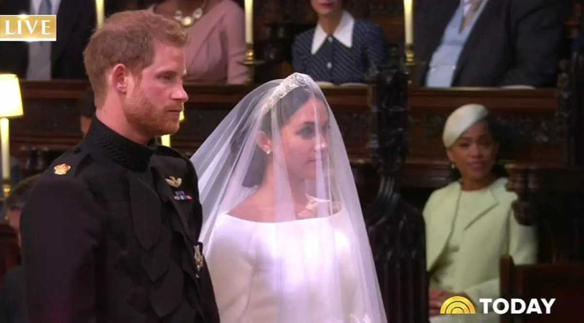 Prințul Harry, purtând o uniformă neagră cu insigne aurii, stând în picioare cu privirea înainte și Meghan Markle, purtând o rochie albă cu umeri goi și un voal subțire care îi acoperă fața și părul.