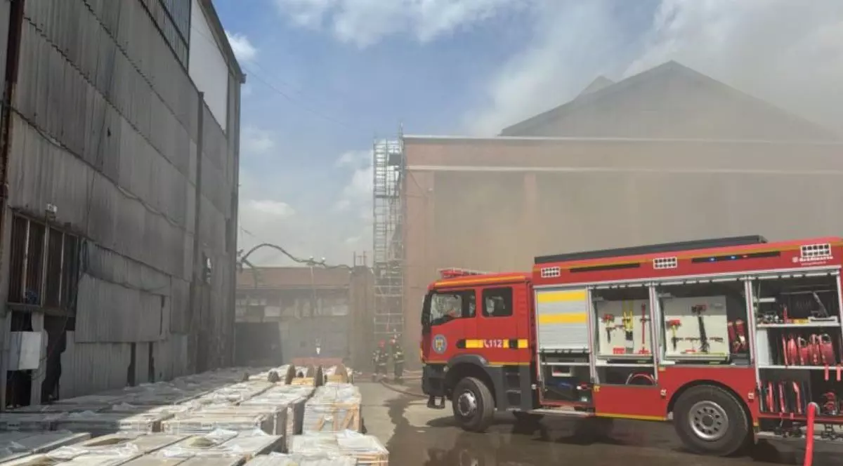 În dreapta o mașină de pompieri lângă o hală cu mult fum în jur