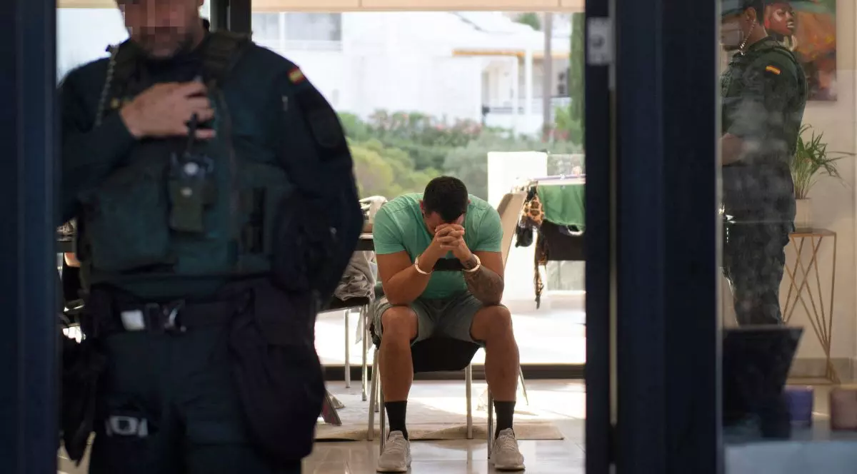 În prim plan un polițist spaniol îmbrăcat în uniformă, iar în plan secund un infractor cu cătușe la mâini, care stă pe un scaun, îmbrăcat în tricou și pantaloni scurți