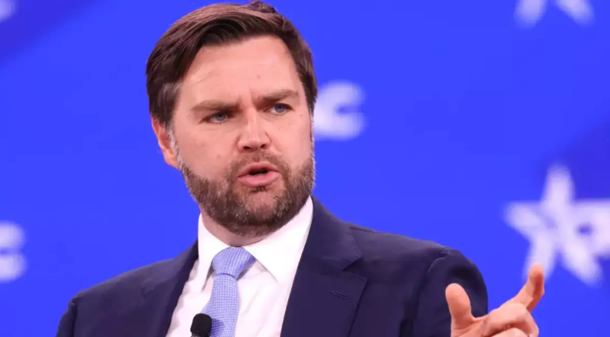 În imagine este ilustrat JD Vance, îmbrăcat în costum albastru închis, cămașă albă și cravată albastră. Acesta are un microfon atașat pe cravată.