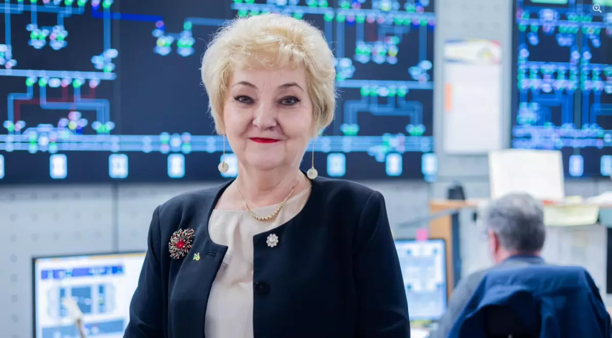 În imagine este o femeie blondă, îmbrăcată în bluză crem, cu un sacou negru. În piept, pe sacou, are o broșă roșie. Aceasta are pe buze ruj roșu și poartă cercei lungi.