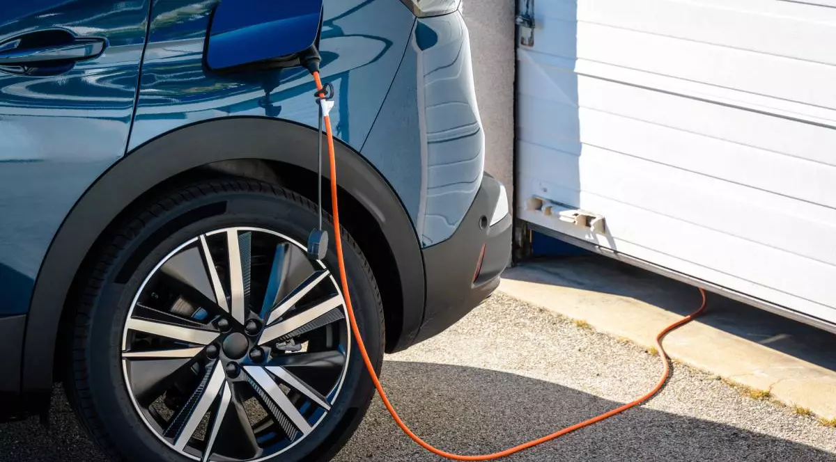 Mașină electrică de culoare albastru închis, parcată lângă o ușă de garaj, conectată pentru încărcare.