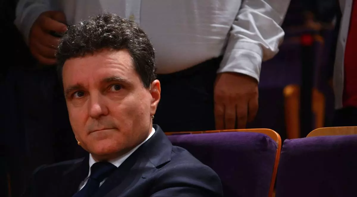 Nicușor Dan, îmbrăcat la costum, în timp ce stă pe scaun într-o sală de evenimente.