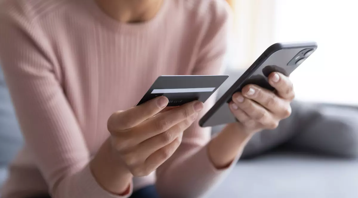 În imagine este ilustrată o femeie care ține în mână un card bancar și un telefon