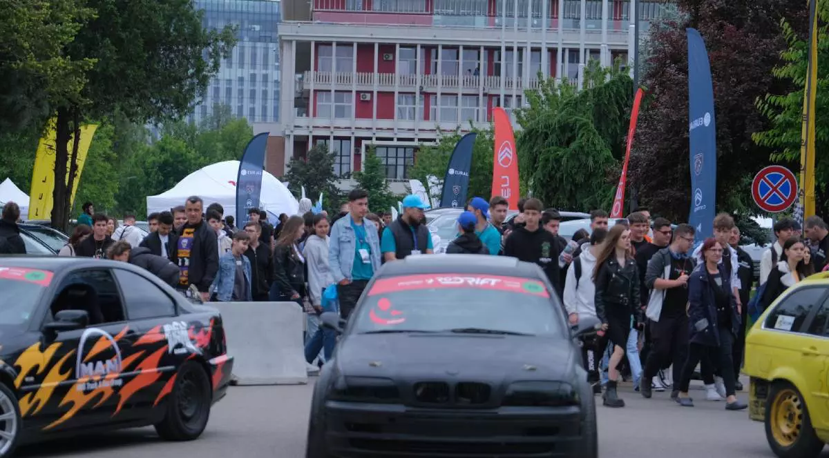 Fotografie din cadrul POLI AutoFest, unde sunt două mașini modificate și un grup numeros de tineri, în mare parte adolescenți și studenți.