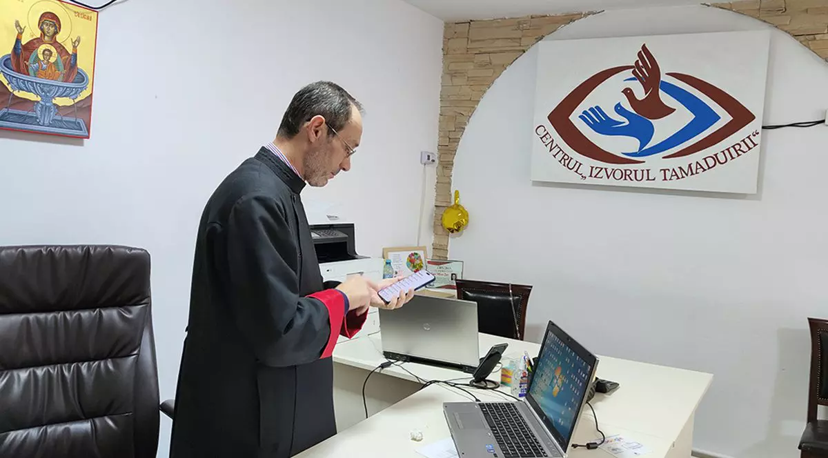 Părintele Liviu Burlacu, centrul Izvorul Tamaduirii, intr-un birou, in fata a doua laptopuri