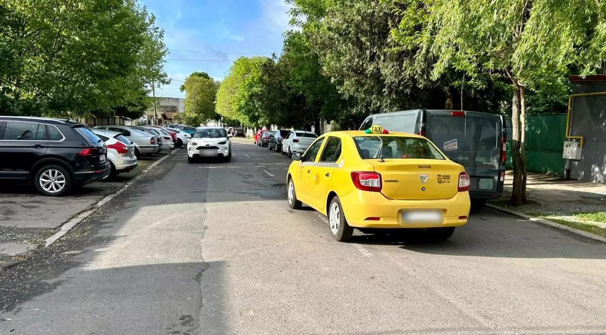 Un taxi galben este cu spatele pe o stradă, iar o mașină albă vine din sens opus, iar în laterale sunt autoturisme parcate, pe o stradă din Constanța