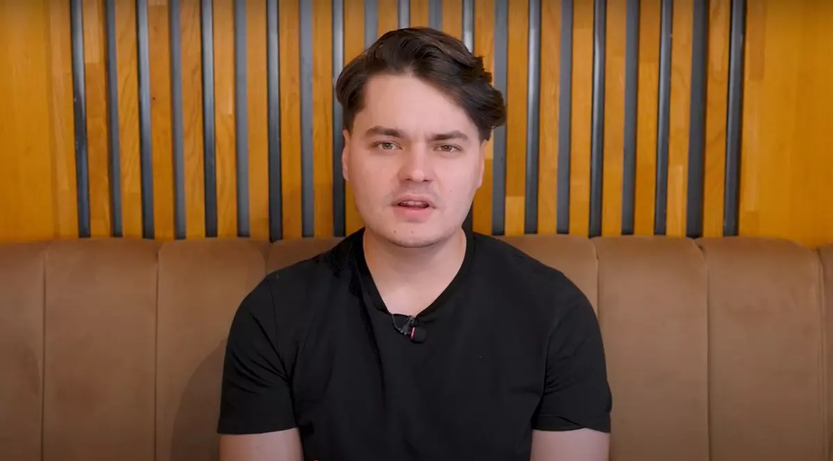 Selly este așezat pe o canapea maro, îmbrăcat într-un tricou negru