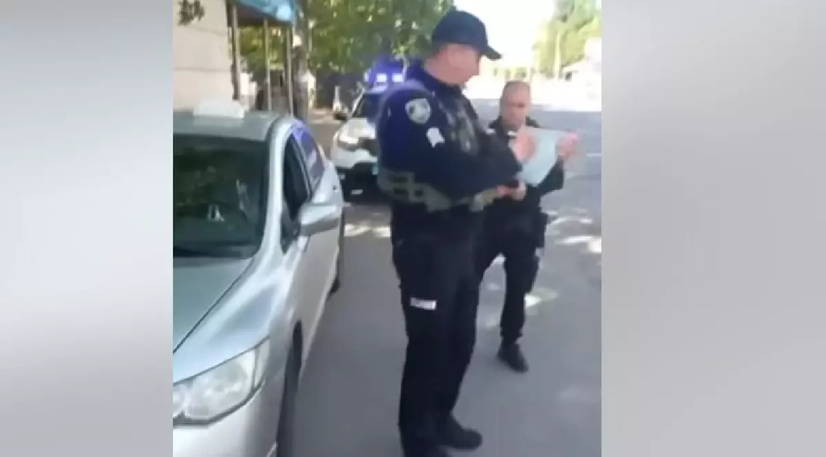 Un bărbat și o femeie, îmbrăcați în uniforme de polițiști, dau amendă unui șofer care a parcat într-un loc interzis. În stânga, o mașină gri parcată pe marginea unei străzi.