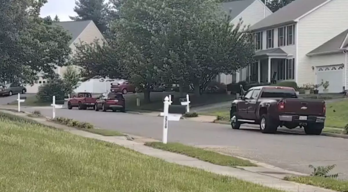 O stradă pe care se văd câteva mașini parcate, case și copaci pe partea dreaptă, iar în stânga este iarbă verde, SUA, mai 2025