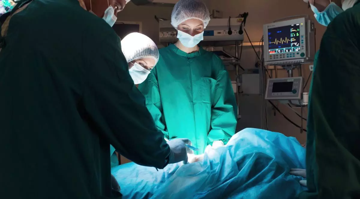 Medici chirurgi, imbracati in costume verzi si cu bonete pe cap, in sala de operatie, aplecati asupra unui pacient