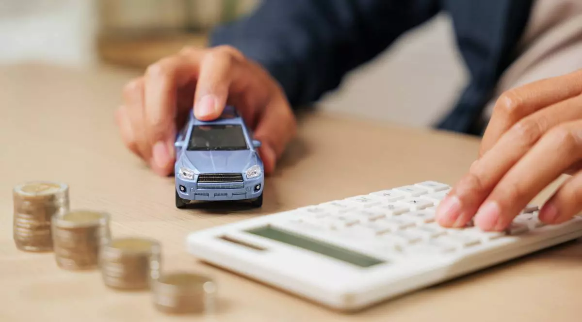 O persoană ține o machetă de mașină albastră într-o mână, lângă este un calculator de birou. Pe masă sunt așezate grămezi de monede, sugerând calcule financiare