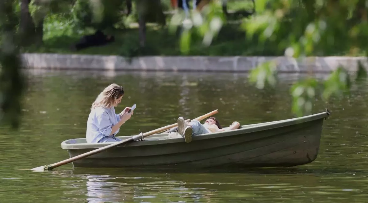 O tânără este într-o barcă pe apă și citește pe telefonul mobil, iar o alta stă culcată în barcă și se vede una dintre vâsle, în parcul Cișmigiu, București, 25 aprilie 2025