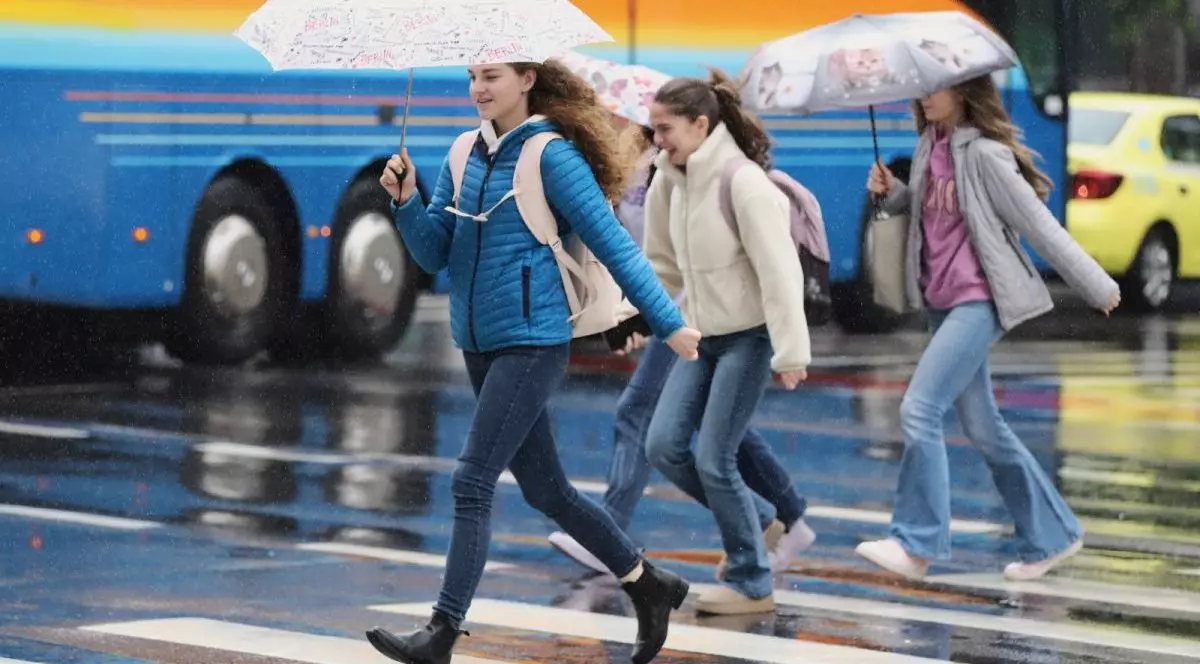 Câteva adolescente, îmbrăcate cu blugi geci, traversează pe trecerea de pietoni cu umbrele, iar în spate se vede un autocar albastru cu portocaliu și un taxi galben, în București, 26 mai 2025