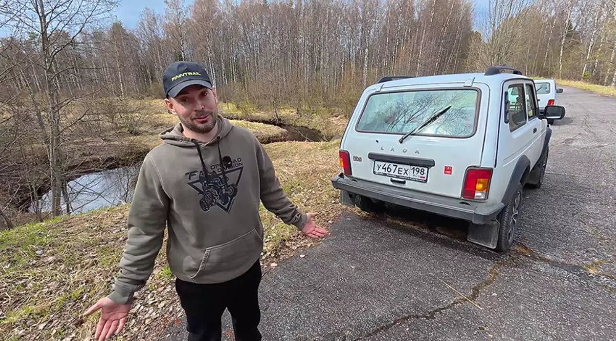 Youtuber rus stând lângă o mașină Lada Niva, parcată pe marginea unui drum de pădure.