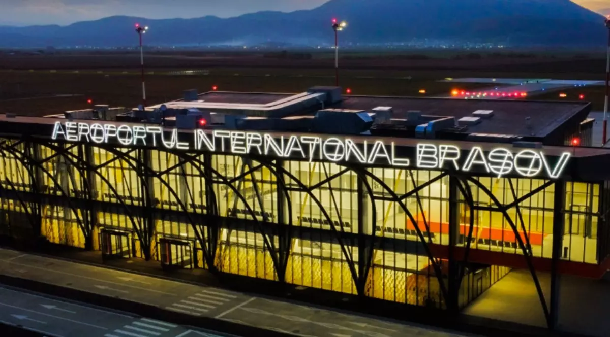 Imaginea prezintă clădirea principală a Aeroportului Internațional Brașov-Ghimbav. Numele aeroportului este afișat proeminent, cu litere mari luminate, vizibile de la distanță.