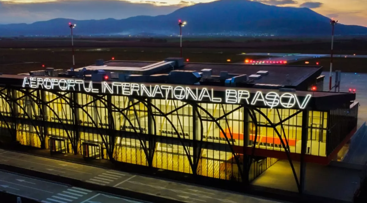 Imaginea prezintă clădirea principală a Aeroportului Internațional Brașov-Ghimbav. Numele aeroportului este afișat proeminent, cu litere mari luminate, vizibile de la distanță.