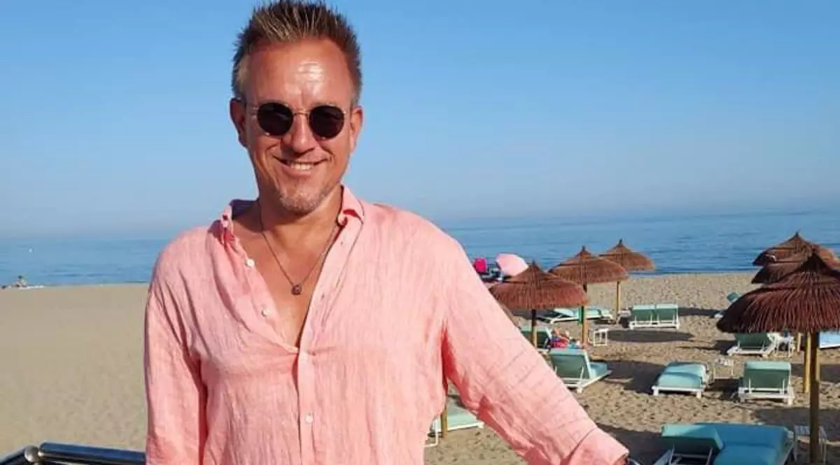 In imagine Anders Kurtén, CEO-ul Fraser Yachts,, intr-o cămașă lejeră roz pudrat, cu ochelari de soare, in spatele lui o plaja cu umbrele de stuf , nisip și marea