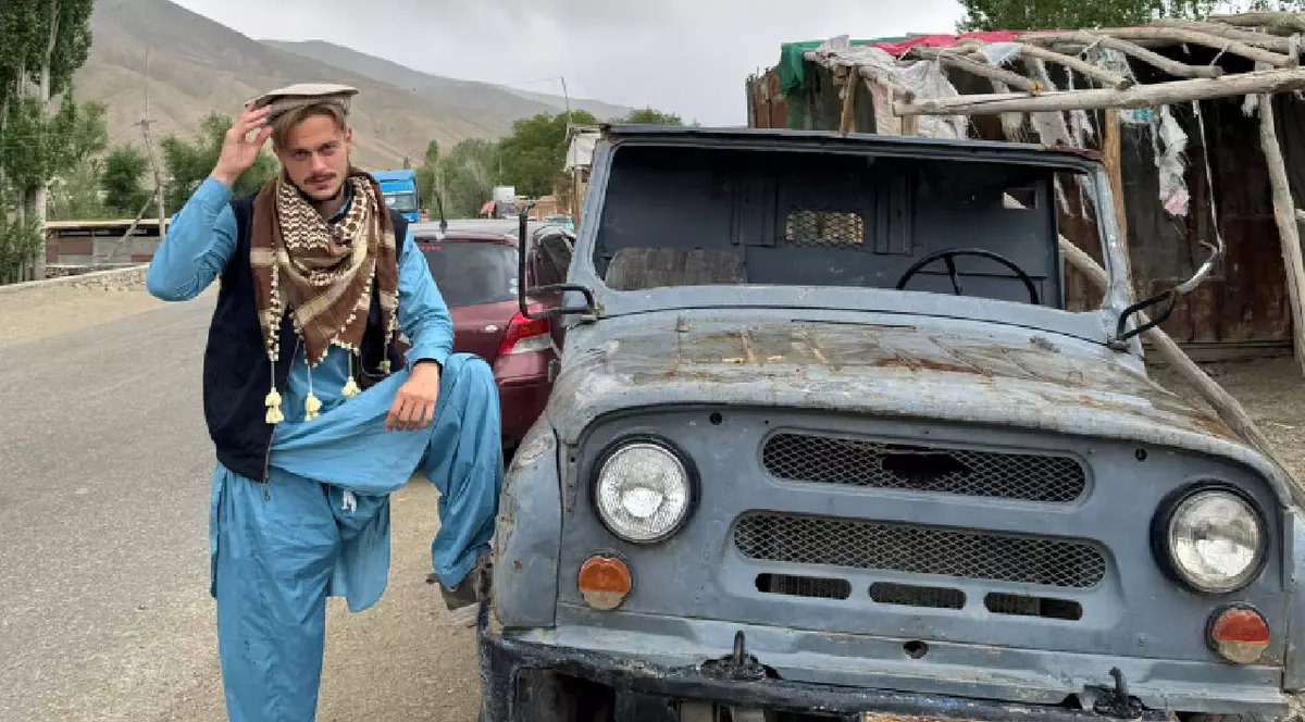 În imagine un bărbat îmbrăcat în haine albastre stă cu piciorul rezemat de mașină pe o stradă din Afganistan