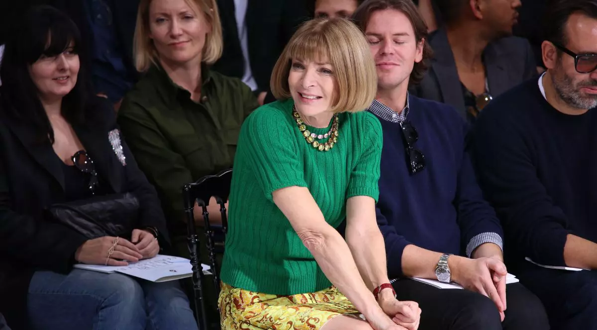 Anna Wintour, așezată în primul rând la un eveniment de modă în Paris, septembrie 2018. Este îmbrăcată un pulover verde cu mâneci scurte, din material tricotat, și o fustă scurtă cu imprimeu floral galben. Stă cu picioarele încrucișate și mâinile împreunate în poală. În jurul ei sunt și alte persoane, care sunt îmbrăcate în culori reci