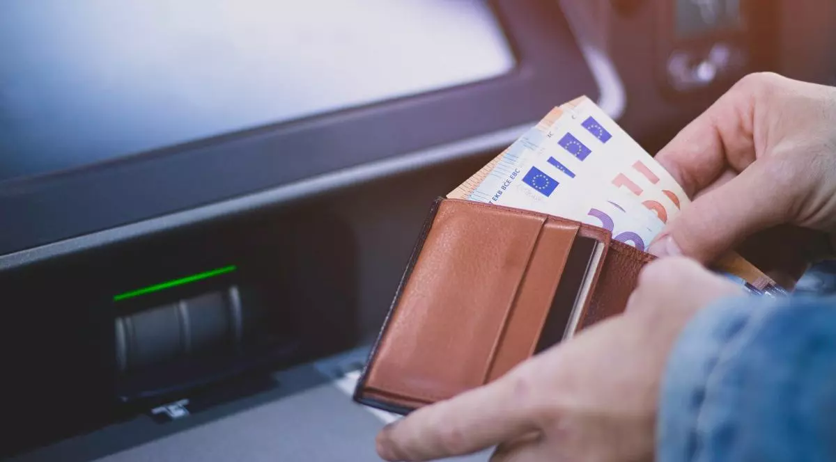 Mâna unui bărbat îmbrăcat cu geacă de blugi care are un portofel din care scoate bancnote de euro