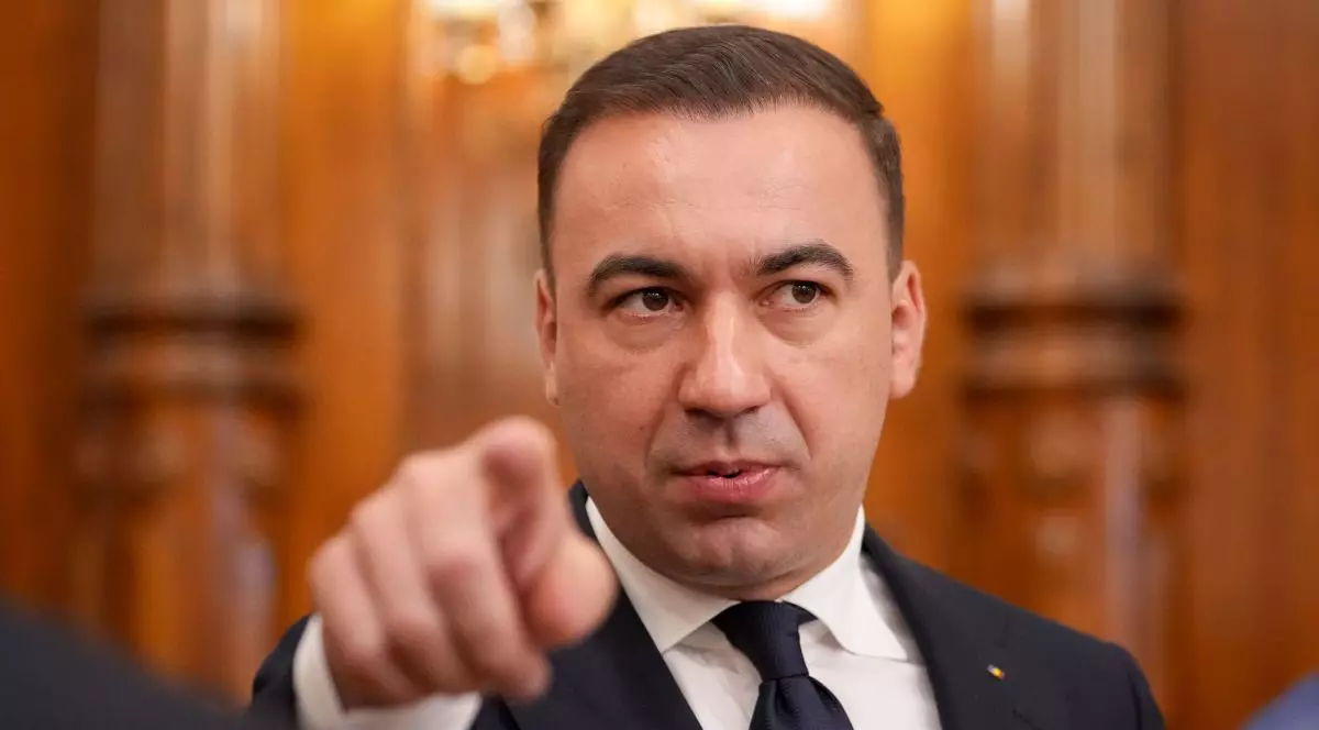 Bogdan Ivan în prim plan, în timp ce arată cu degetul către cameră, îmbrăcat într-un costum negru, cămașă albă și cravată neagră