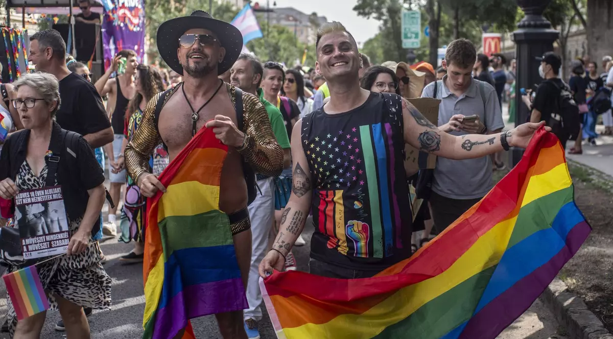 Imagine surprinsă în timpul Budapest Pride din iunie 2024. Doi bărbați se află în prim-plan, iar ambii țin în mâini câte un steag LGBTQ. Cel din stângă nu poartă tricou, are o pălărie și ochelari de soare. În spatele lor se află mai mulți oameni