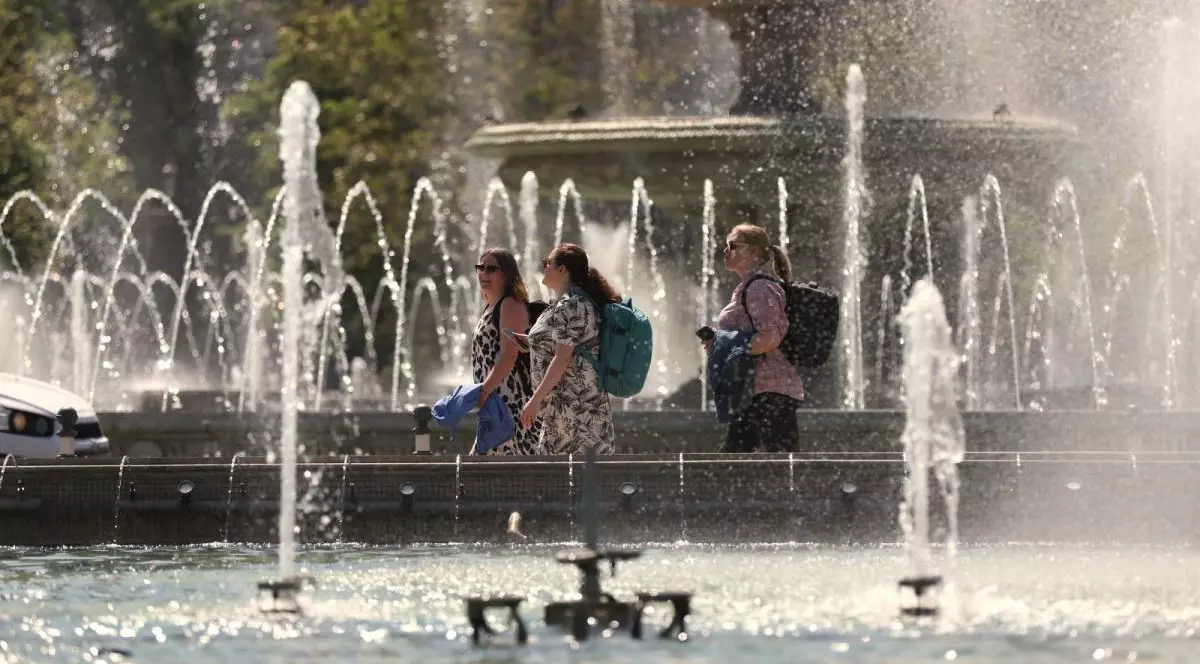 Trei femei cu ghiozdane în spate trec prin dreptul unei fântâni arteziene, unde se vede apa țâșnind, într-o zi foarte caldă, București, 24 aprilie 2025
