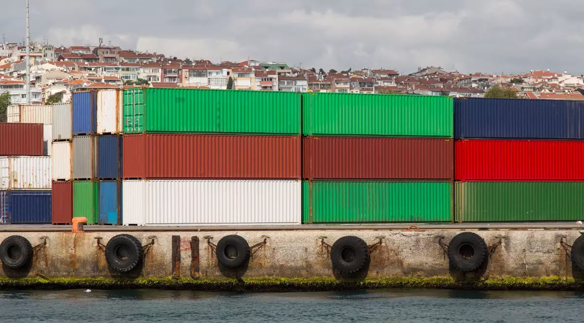 containere metalice de marfă de culori diferite: verde, roșu și alb, așezate unele peste altele, într-un port. pe marginea docului sunt prinse cauciucuri de mașină și în fundal se văd case în depărtare cu acoperișuri portocalii și pereți albi șo crem