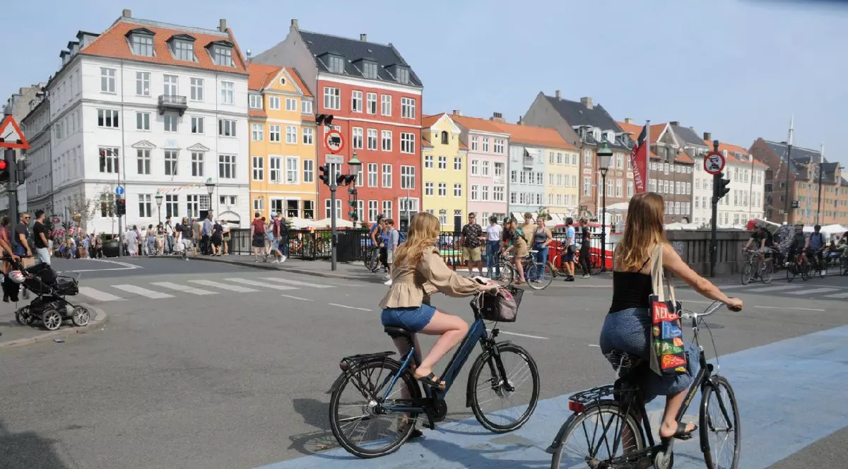 Două fete merg pe biciclete în fața unor case colorate din Copenhaga, in jurul or sunt persoane care se plimba