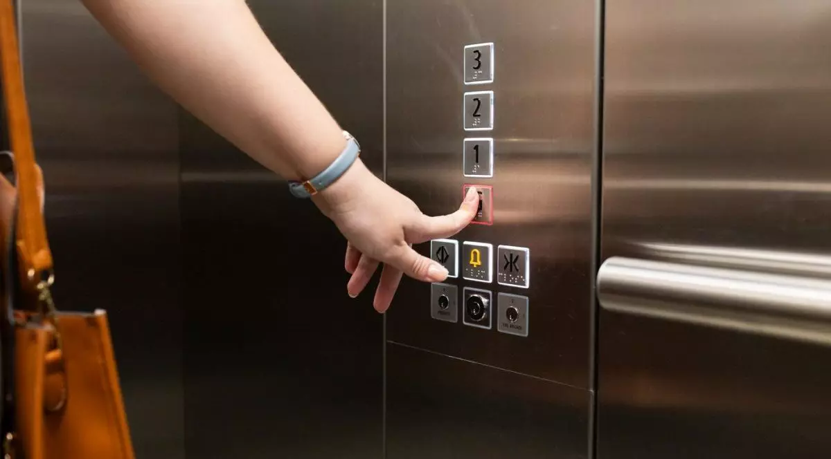 Mâna unei femei cu ceas gri și o geantă maro pe umăr care apasă cu degetul pe butonul unui lift în care se află