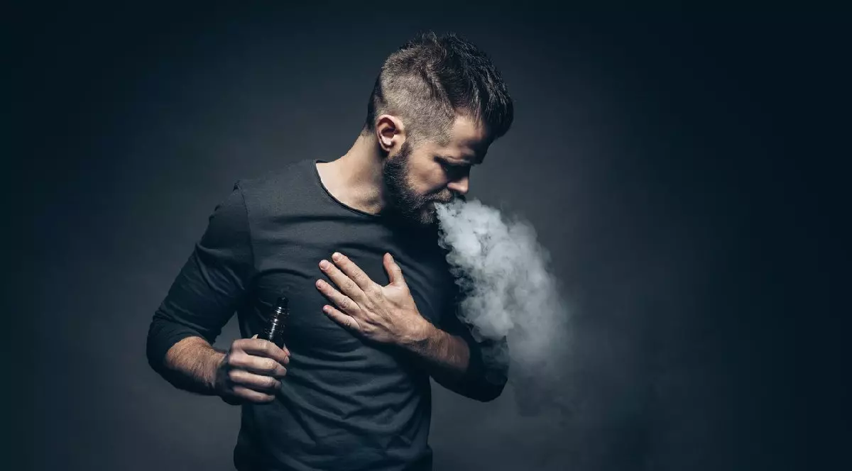 Un tânăr îmbrăcat în bluză neagră, pe fundal negru, vapează dintr-o tigară electronică- Vaporizarea e-lichidului aromatizat de la o țigară electronică