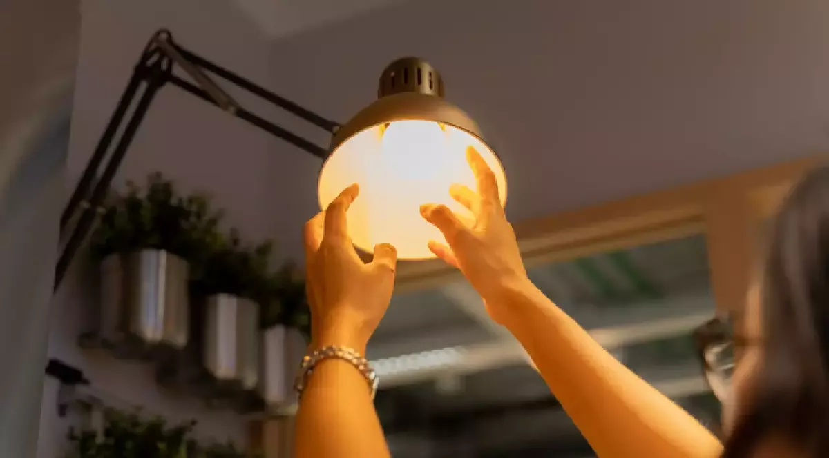 O femeie înșurubează un bec cu mâinile într-o veioză și se vede lumina