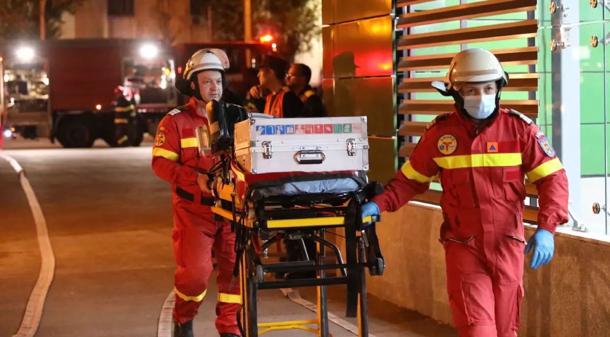 In imagine doi salvatori SMURD înbrăcați în combinezoane rosii transporta echipament pe o targă, seara, în cadrul Exercițiului SEISM 2025 Convex 3