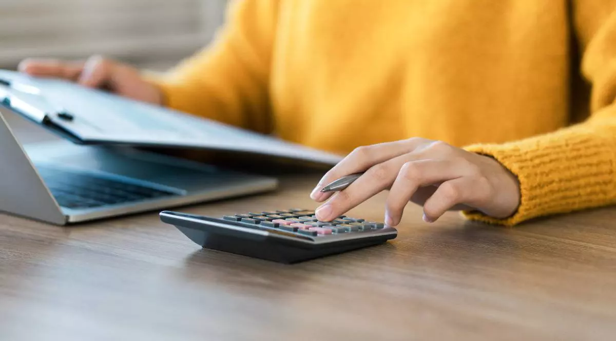 O femeie care stă în fața calculatorului, ține o serie de hârtii în mână și face calcule pe un mic calculator manual.