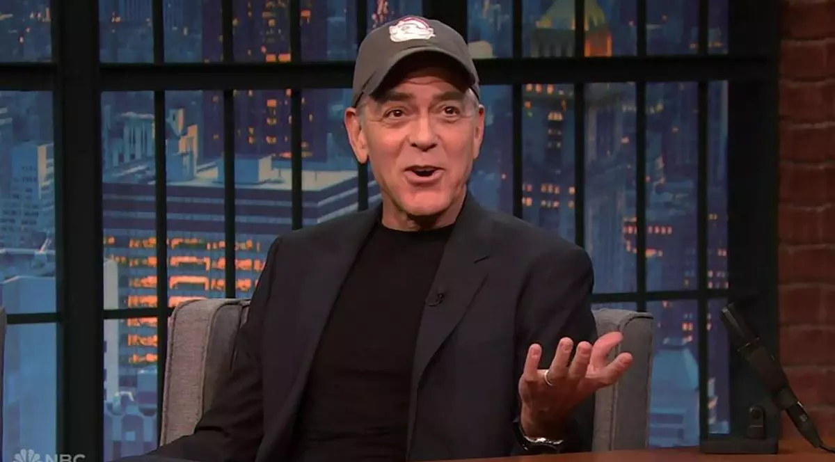 George Clooney în timpul unei emisiuni TV, stă pe canapea într-un costum negru, gesticulează cu mâinile și poartă o șapcă pe cap.