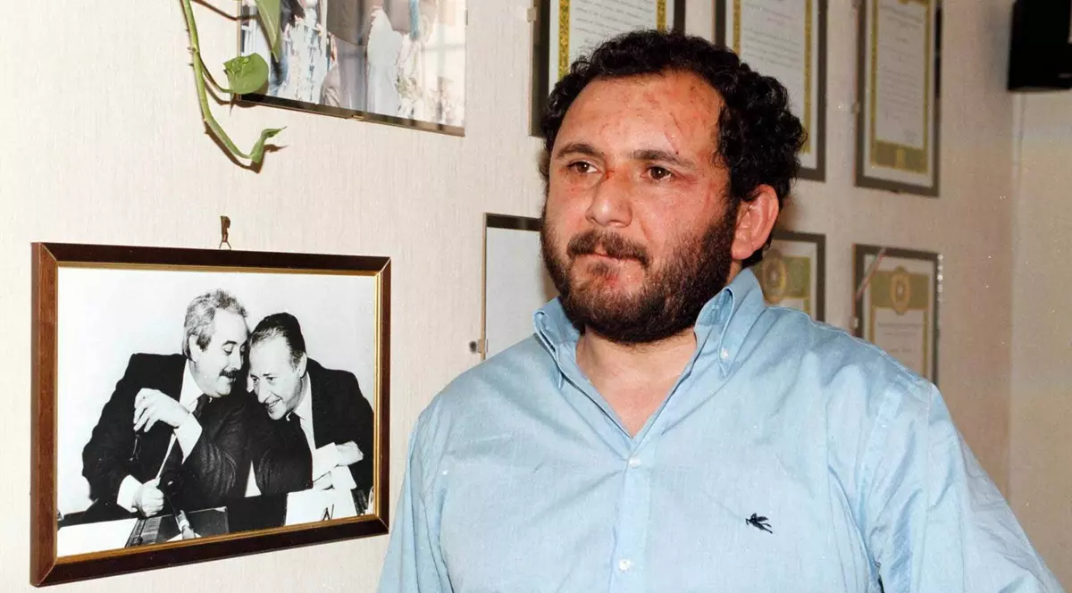 Giovanni Brusca poartă o cămașă albastră, iar în spatele său sunt atârnate pe perete fotografii