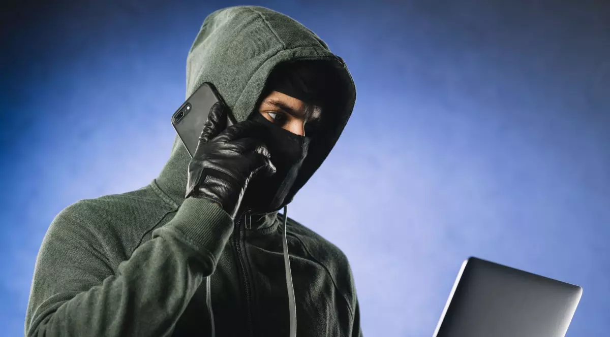 Un tânăr în hanorac verde cu gluga trasă pe cap și cu o cagulă pe față vorbește la telefonul mobil în timp ce se uită pe un laptop