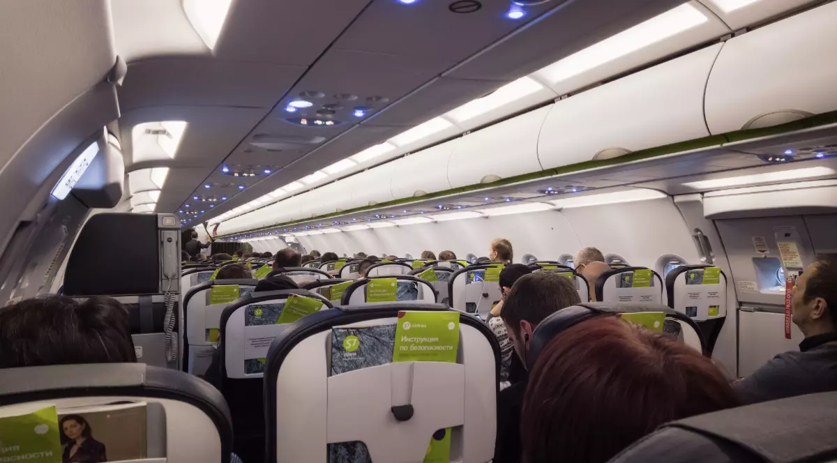 În imagine este ilustrat interiorul unui avion în care pasagerii se află pe scaune.