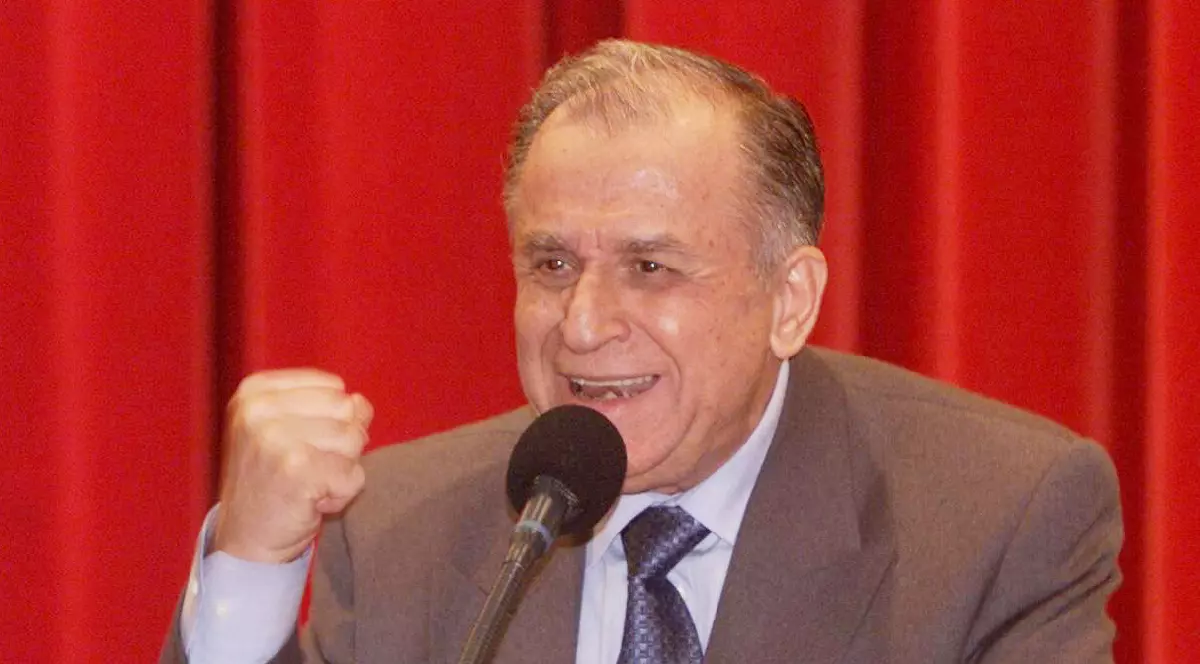 Ion Iliescu în costum vorbind la microfon, cu pumnul strâns, pe fundal roșu