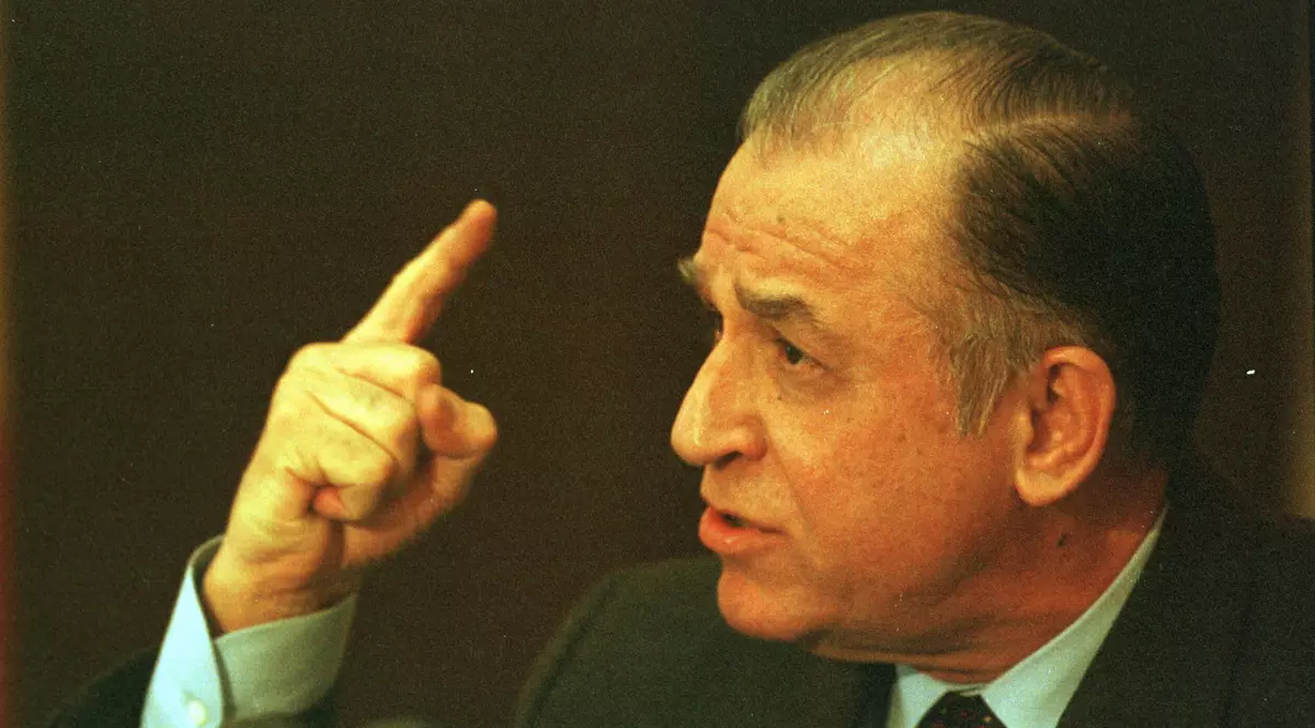 Ion Iliescu surprins în timp ce ține un deget ridicat. Este îmbrăcat cu un sacou închis la culoare, poartă o cravată roșie și o cămașă bleu