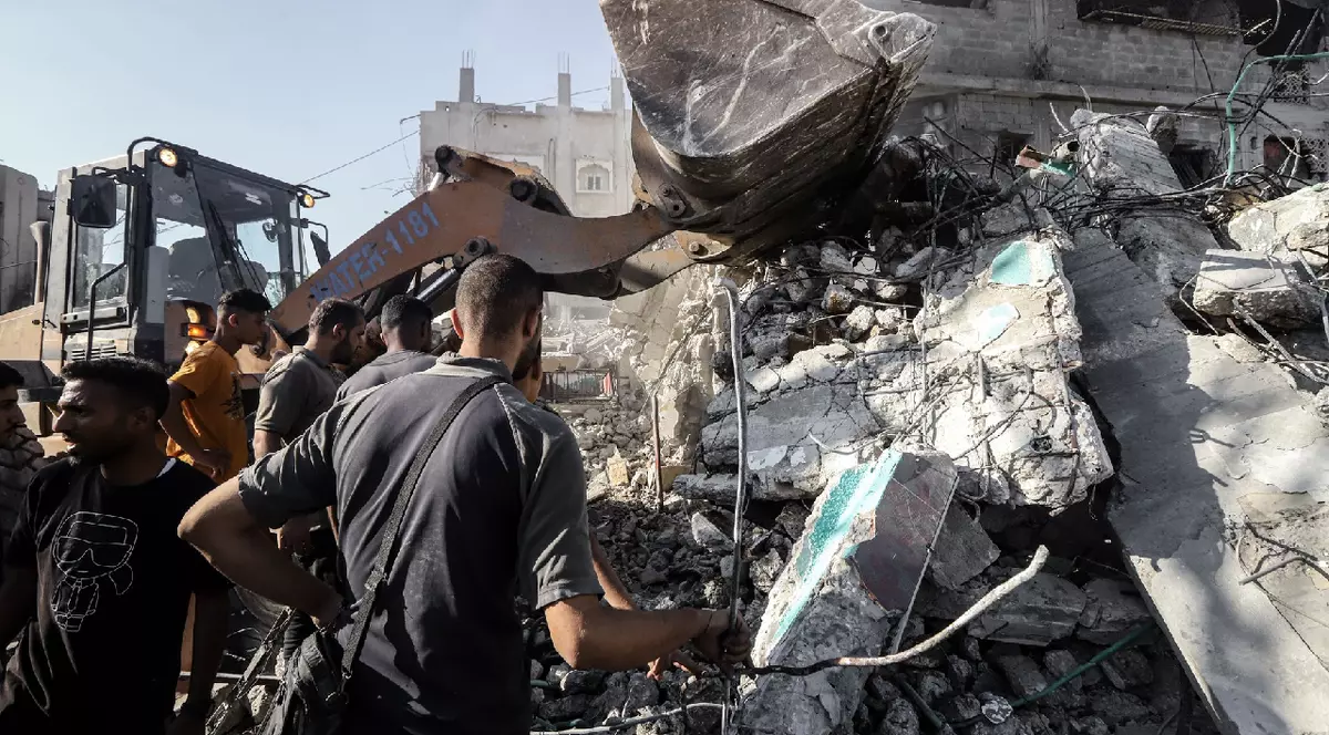 Mai mulți bărbați inspectează ruinele unei clădiri distruse, în timp ce un excavator încearcă să degajeze dărâmăturile într-o zonă afectată de conflict