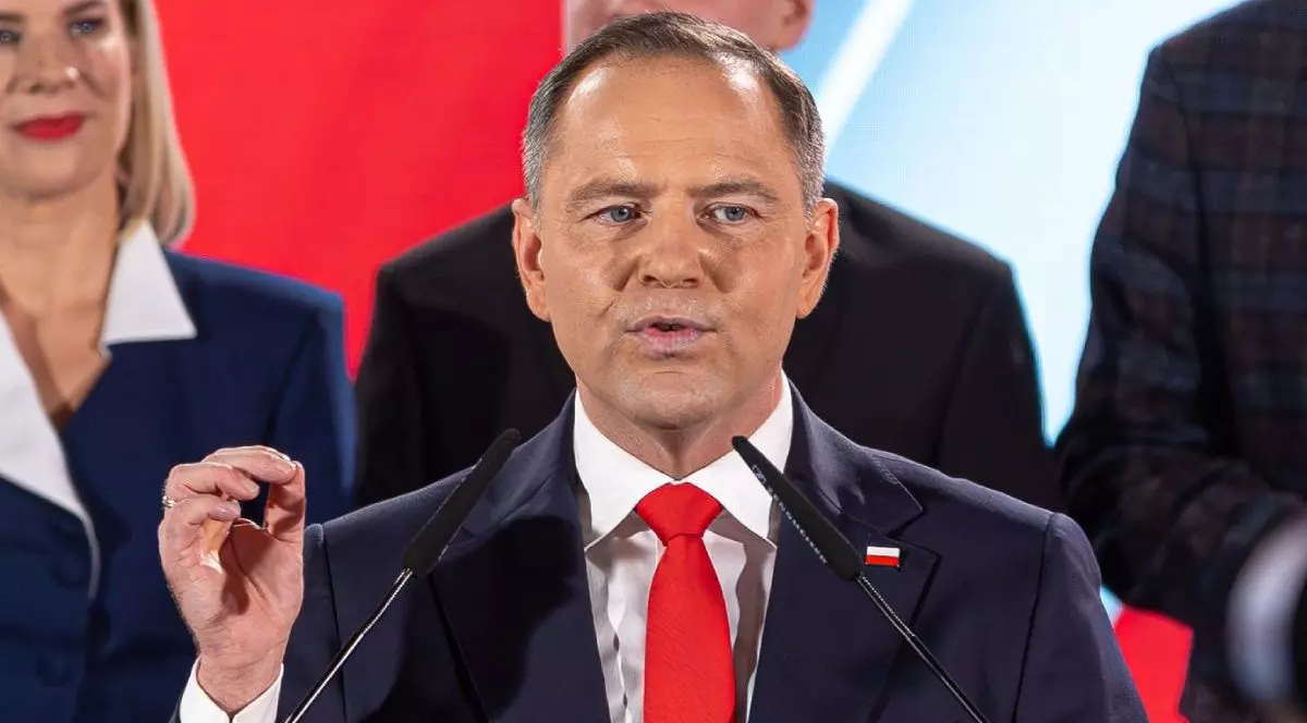 Karol Nawrocki, in sacou negru si cravata rosie, vorbeste la microfon cu mana dreaptă ridicată si oameni din staff în spate