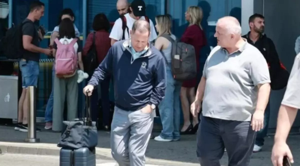 În prim-plan se află doi bărbați: unul cu un troller și o geantă de mână, îmbrăcat într-o jachetă sport și pantaloni deschiși la culoare, care a aterizat recent; celălalt poartă un tricou gri și pantaloni închiși la culoare, și îl privește pe primul.