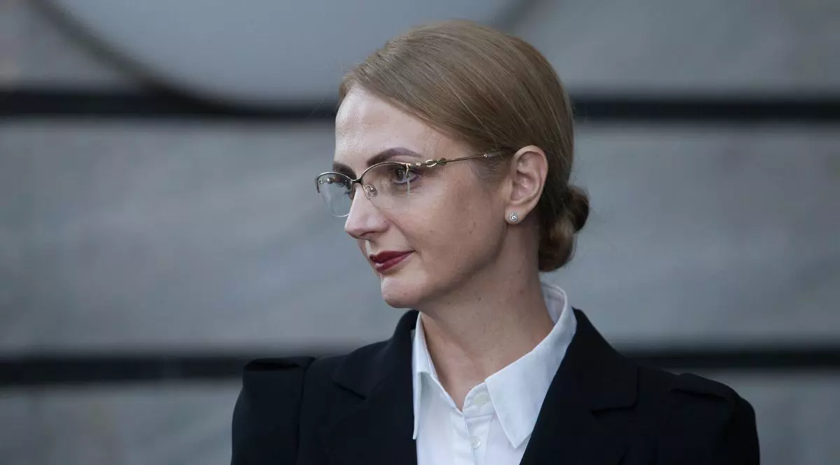 Lia Savonea, fotografiată din semi-profil, purtând un sacou negru și o cămașă albă, cu un coc jos simplu, ochelari rotunzi cu ramă aurie și ruj roșu pe buze