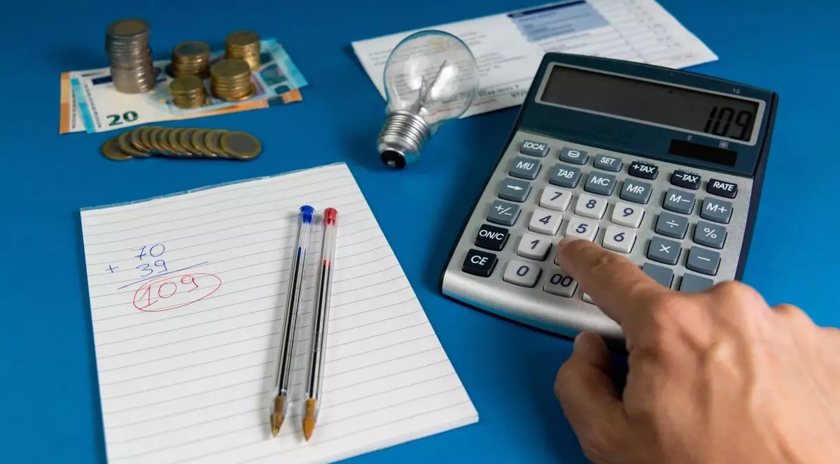 O factură de electricitate cu un bec, o mână calculează factura folosind un calculator și un caiet cu două pixuri și multe monede euro pe fundal albastru, iar sub ele este o hârtie cu un calcul și două pixuri, unul albastru și unul roșu