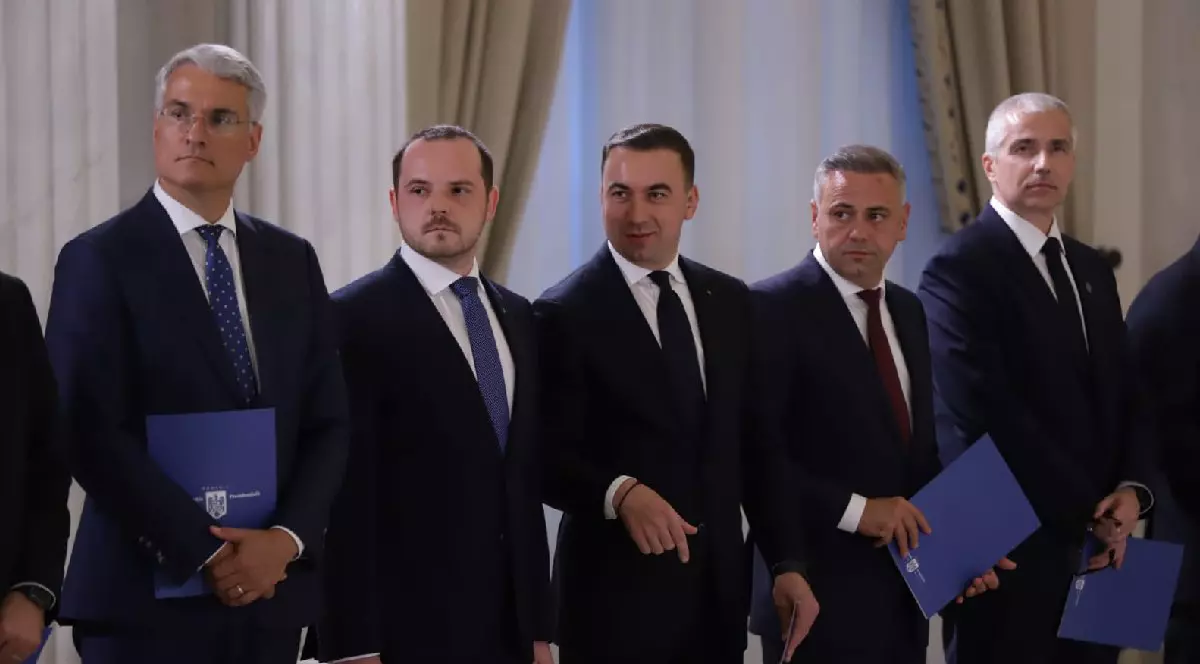Miniștrii din Guvernul Bolojan, îmbrăcați în costume negre, țin o mapă albastră în mână, sunt aliniați pentru o poză.